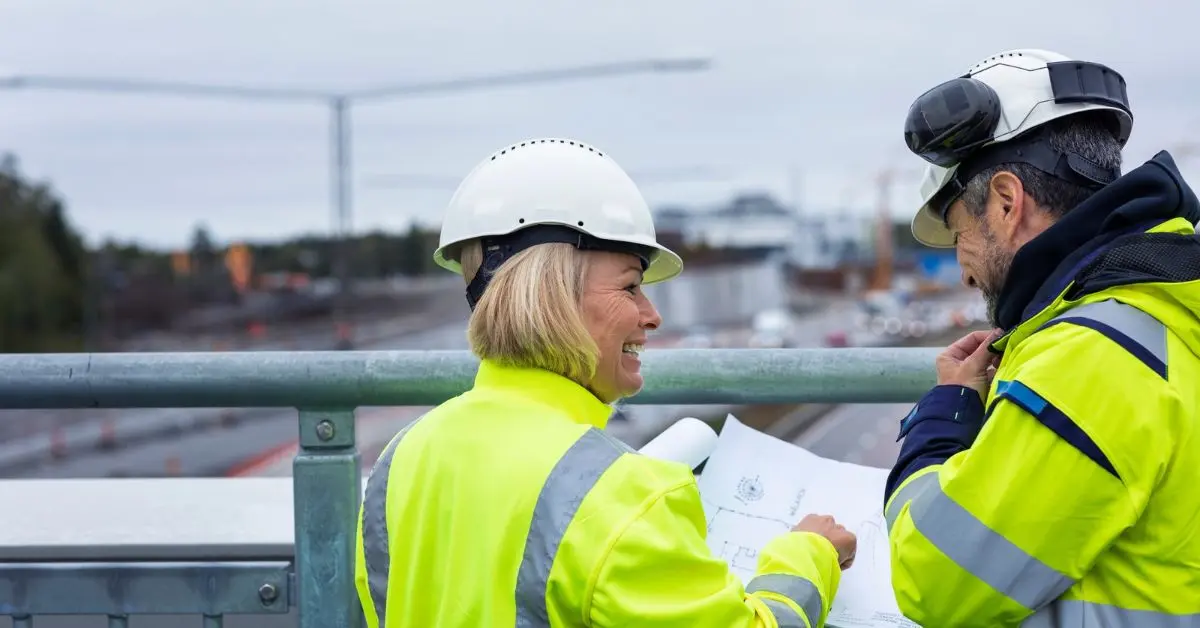Två medelålders byggarbetare diskuterar en skiss i utomhusmiljö.