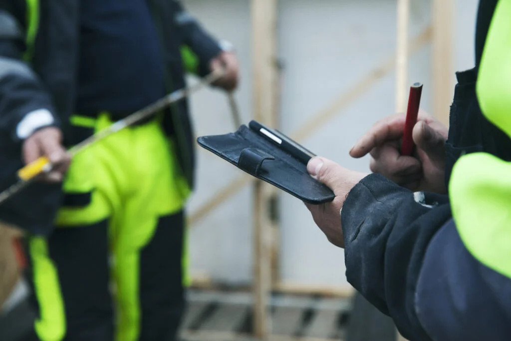 Personer med varselkläder på byggarbetsplats där en ena kollar sin mobiltelefon och den andra håller i mätverktyg